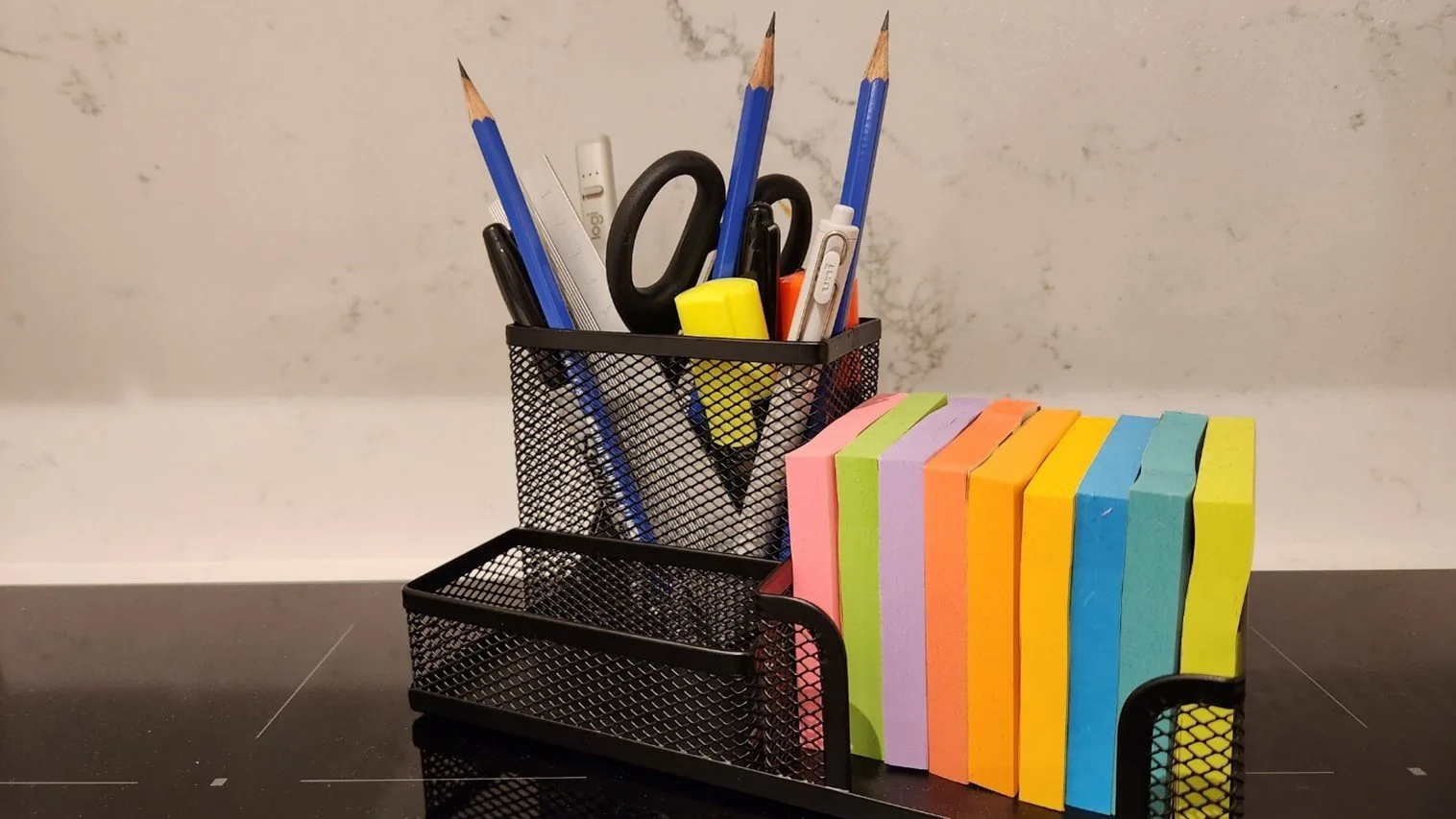 Mesh desk organiser with three pencils, pens, a yellow highlighter, scissors and a ruler, next to a row of coloured sticky note pads