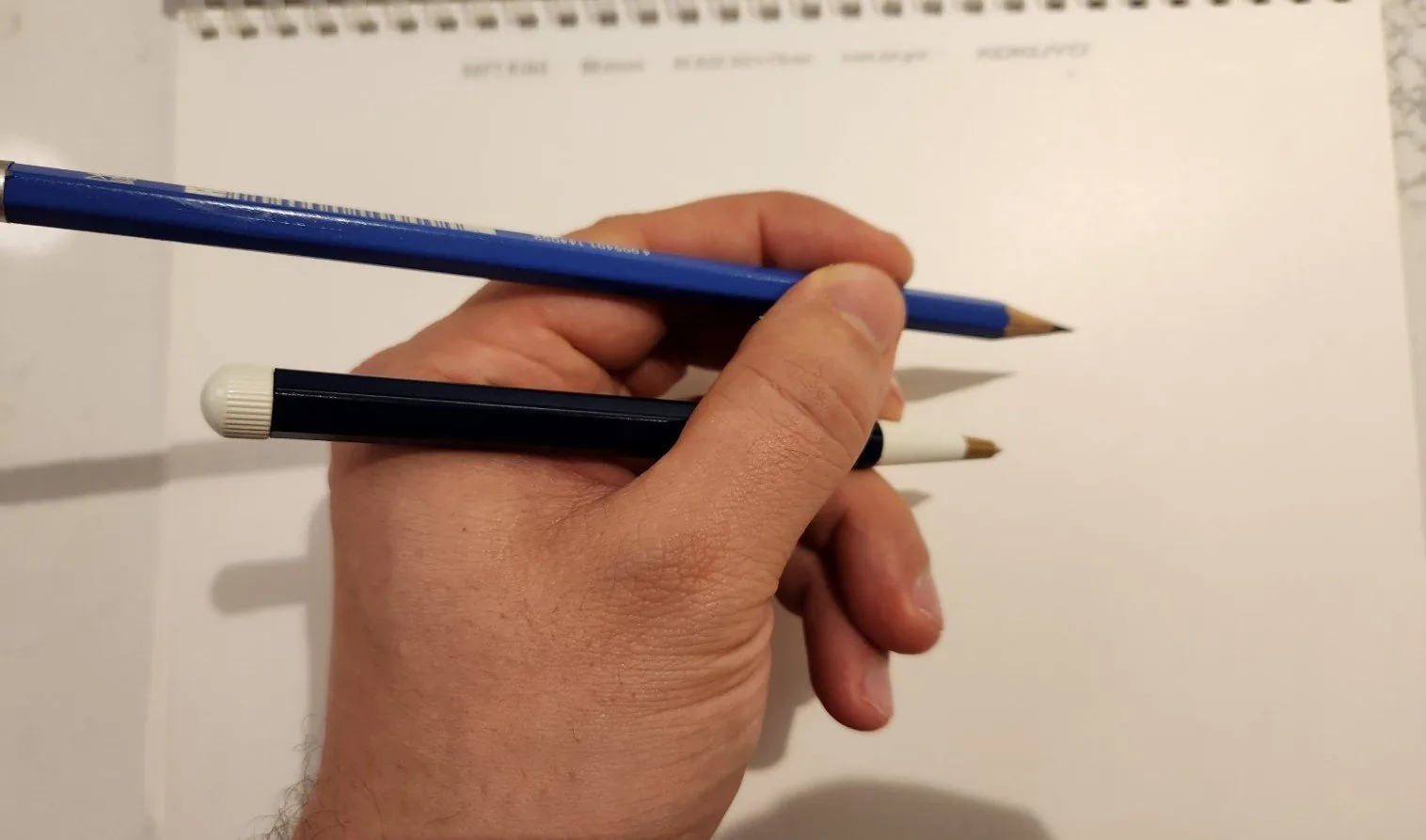 A hand holding two writing tools at once between the fingers: a blue pencil and a capped dark blue pen, over a blank notepad