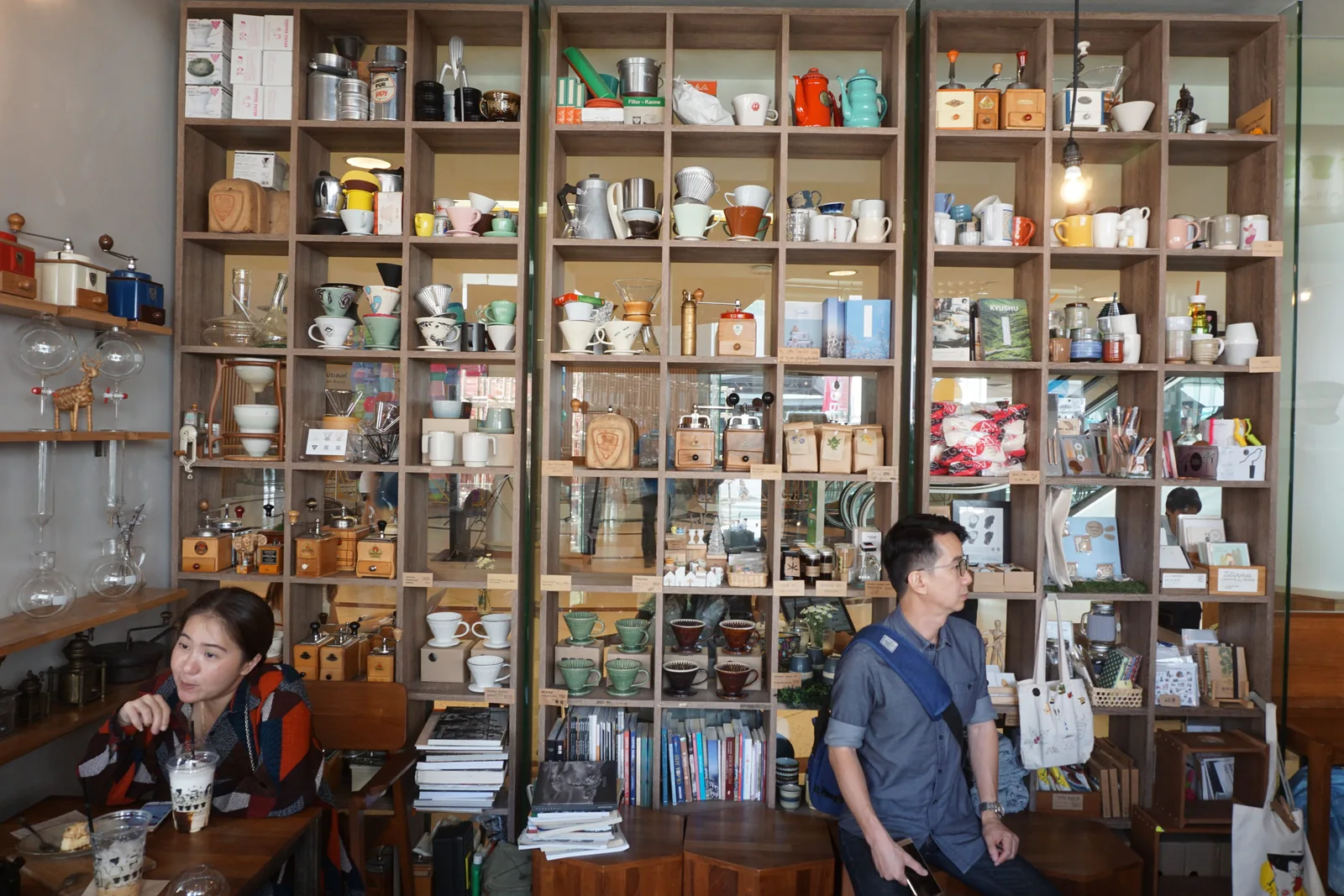 Two guests sitting in front of a floor-to-ceiling shelf of cups, jars and coffee gear