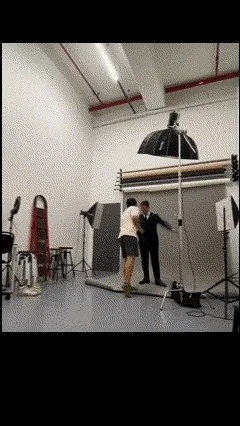 A photo studio: the subject in a suit stands against a backdrop while the photographer adjusts the pose, softboxes and an octabox around