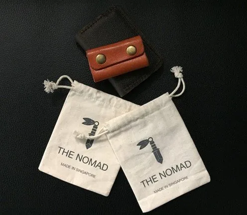 Два хлопковых мешочка с принтом THE NOMAD и подписью MADE IN SINGAPORE, рядом коричневый кожаный кабель-холдер